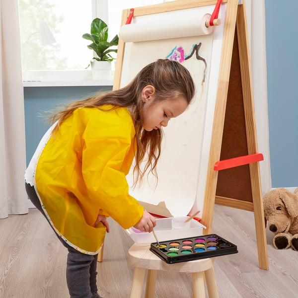 A child in a yellow apron stands at a wooden easel, painting with watercolours.