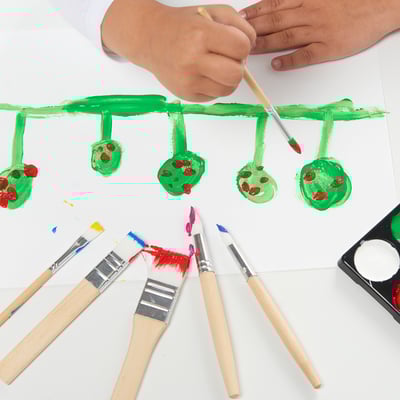 Childs hands painting green leaves and red spots on paper with assorted brushes.