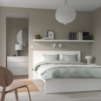 Modern bedroom with a white MALM bed centred, small chair left, floating shelf above with books, lamp, and art, large mirror in background.
