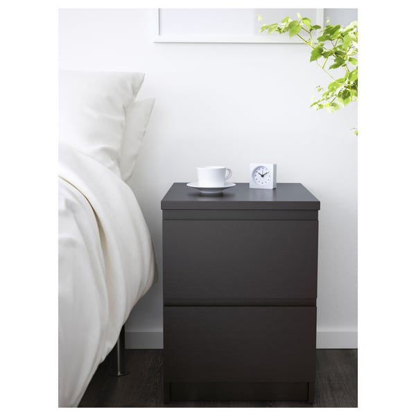 Modern dark grey MALM bedside table beside white bed, holding white mug, plate, and clock, with green leafy plant above.