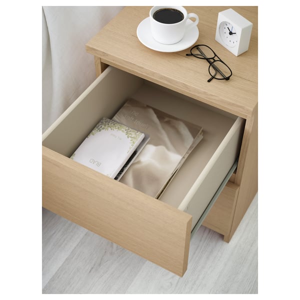 A wooden bedside table with an open drawer holding magazines and a book. On top, theres a cup of coffee, glasses, and a clock.