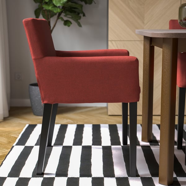 Red upholstered chair with armrests and dark legs on striped rug by wooden table.