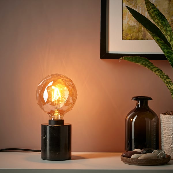 Modern desk lamp with black marble base and lit bulb beside a vase and framed plant picture.