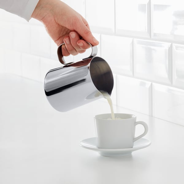 Hand pouring frothing milk jug into white coffee cup on saucer, demonstrating milk frothing.