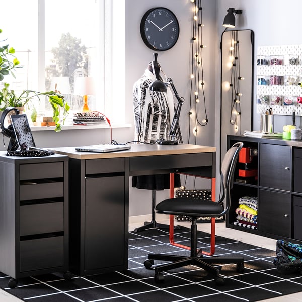 Modern home office with black MICKE desk, red chair, clock, plant, and string lights.