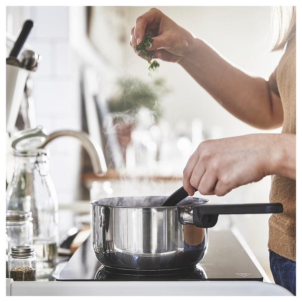 Person cooking in a modern kitchen, sprinkling herbs into a stainless steel MIDDAGSMAT saucepan on a stove.