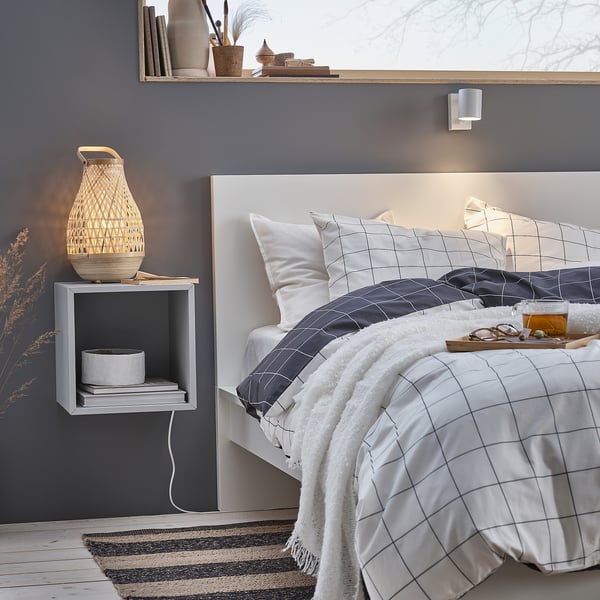 This image shows a serene bedroom with a white MISTERHULT bamboo lamp on a grey bedside table. The bed has white and grey grid-patterned bedding.