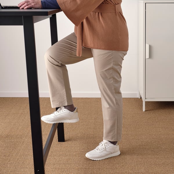 Person leaning on high table, using footrest for support. Table has hooks and cable management. Ideal for meetings or work.