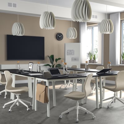 Modern office conference room with MITTZON tables, beige chairs, white pendant lights, black tv, and clock. Tables large, with laptops and cups.