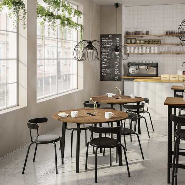 Modern cafe with wooden tables, black chairs, plants, large windows, and a blackboard menu.