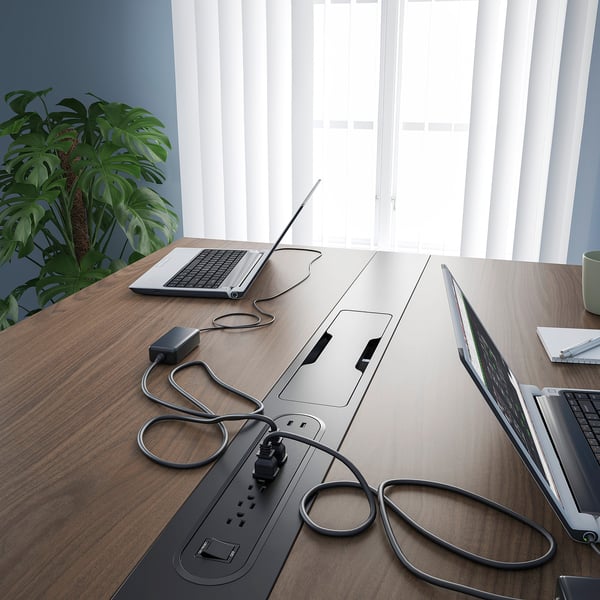 Wooden conference table with integrated power strips and open laptops, near a window with blinds and a potted plant.