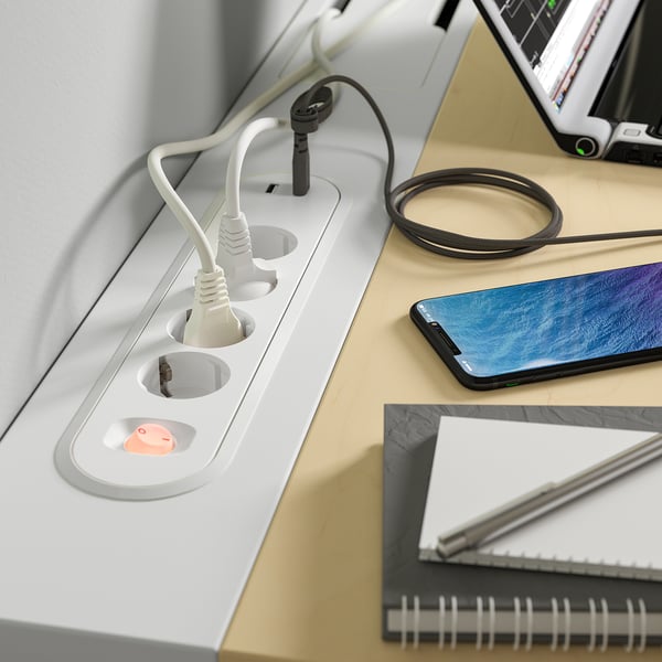 White desk with built-in power strips and cords; smartphone, notebook, and pen on top.