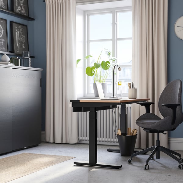 Home office with MITTZON desk, sleek black with wooden top, standing tall beside window, cradling laptop, plants, and chair.