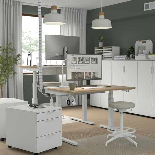 Modern home office with MITTZON sit-stand desk, ergonomic stool, lamps, file cabinet, and computer setup.