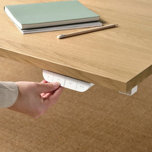 Hand adjusts sit-stand desk button, control panel visible, books and pencil on desk.