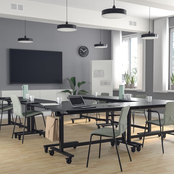 Modern office with mitzon black foldable tables and light green läktare chairs. Large tv, grey walls, big windows. Tables have hooks for bags.
