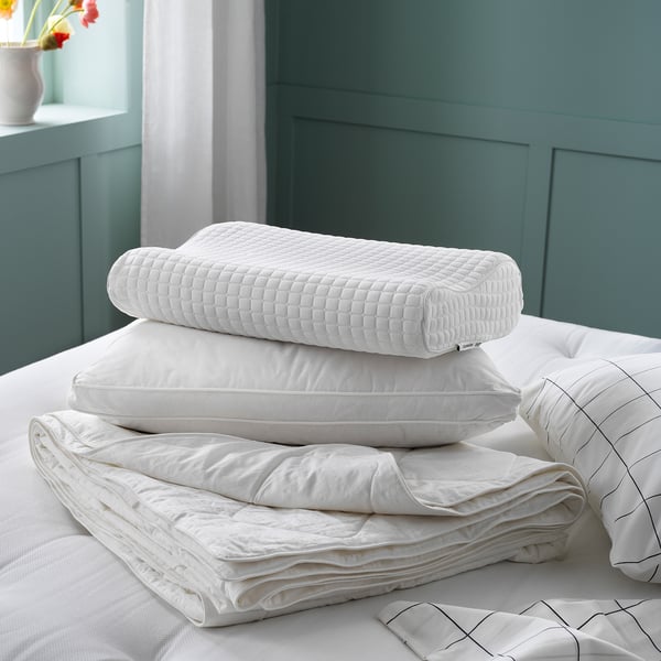 Three stacked white pillows on a bed. One has a quilted design, one smooth cotton, and folded silk. Against teal wall.