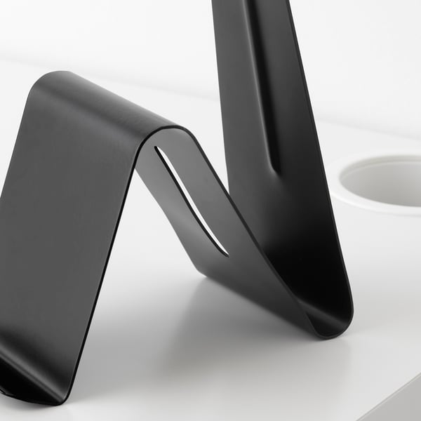 Black metal stand with smooth, rounded edges; holds tablets or phones, sits on white desk.