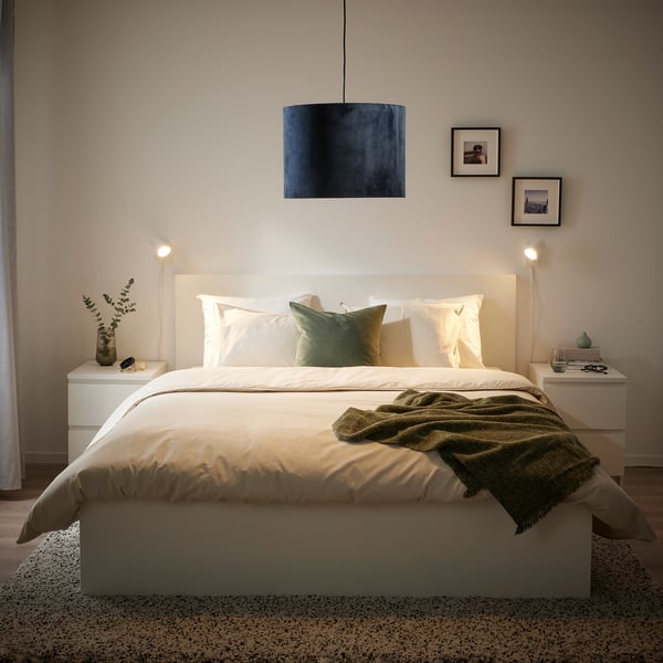 Modern bedroom with MOLNSKIKT lamp. Dark blue, recycled polyester shade glows warmly above white bed with green accents, large and circular.