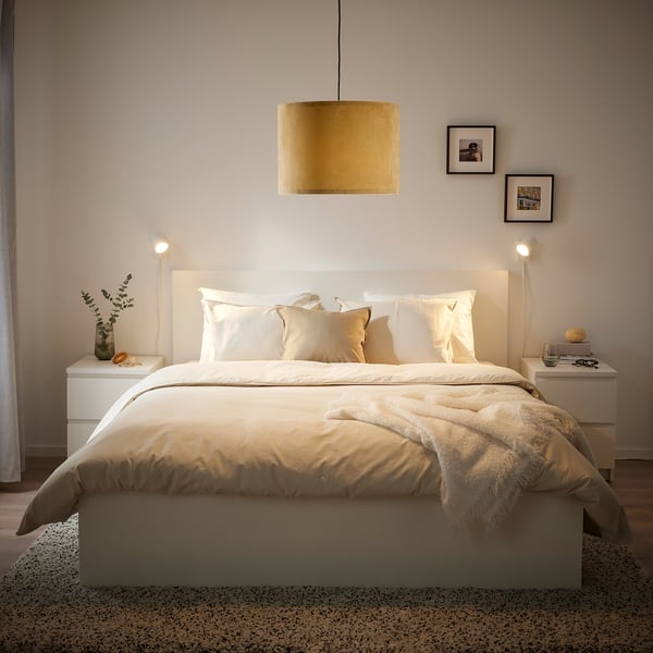 A modern bedroom with a large white bed, yellow velvet lampshade, and white nightstands with vases and nightlights, framed prints on the wall.