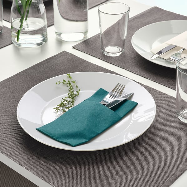Grey table setting with white plate, green napkin, silverware, and glass cup.