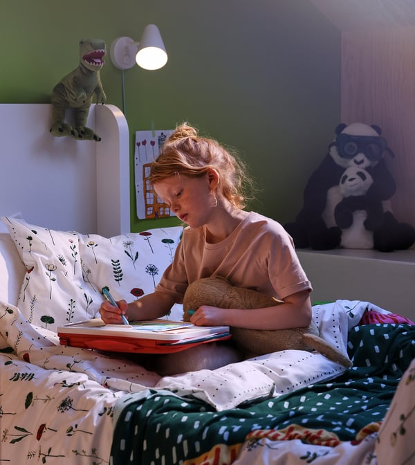 Girl drawing on bed with NATTSLÄNDA duvet, teddy, lamp, panda.