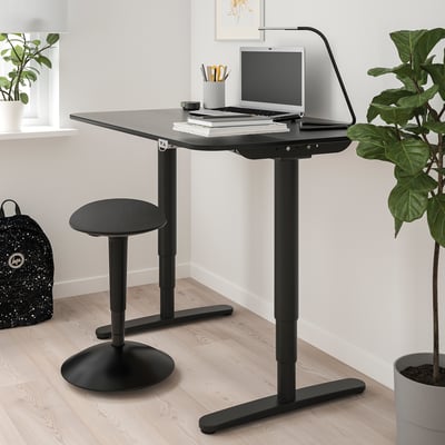 Modern black home office setup with height-adjustable desk & stool, laptop, books, lamp & green plants.
