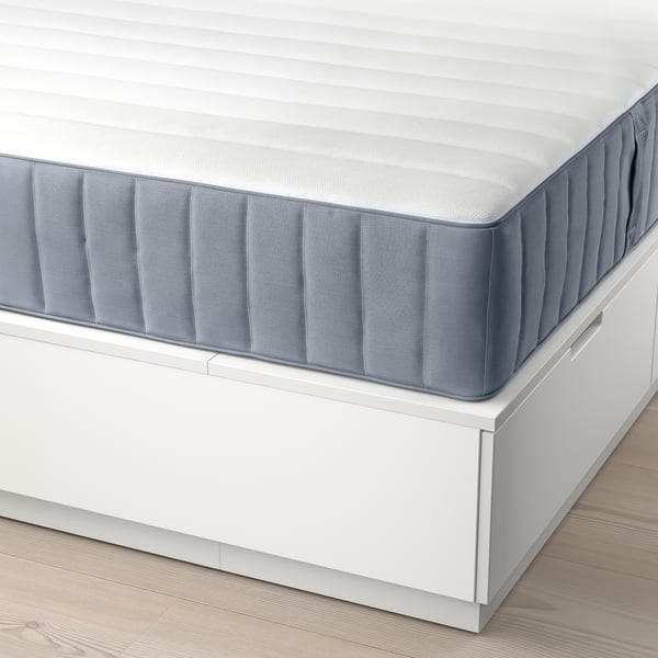 Corner view of a white mattress on a wood floor. Mattress has grey edge cover, resting on white bed base with drawers.