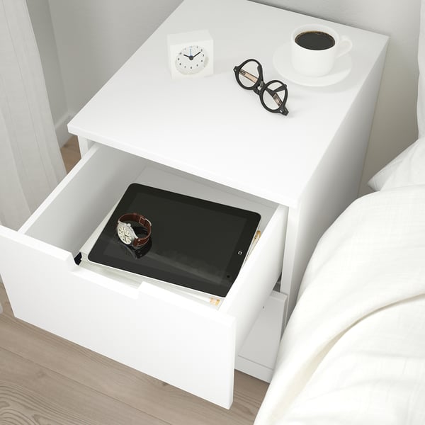 Bedside table with drawer open showing tablet and watch. Clock, glasses, and coffee on top.