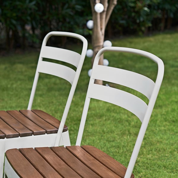Two white NORRMANSÖ metal chairs with wooden seats outdoors. Ideal for dining, light, sturdy, with curved backs.