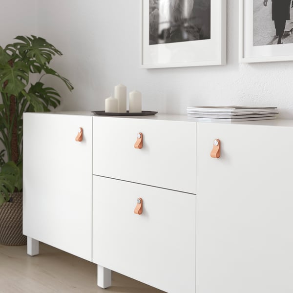White cabinet with leather handles; three drawers, one top shelf with candles and magazines.