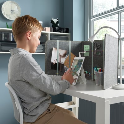 Boy sitting at white desk using tablet. Desk has grey divider for privacy and sound dampening; holds notes and supplies.