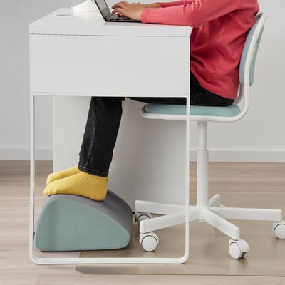 Person in pink top using white desk, feet on blue footrest. The footrest supports correct sitting posture and reduces pressure.