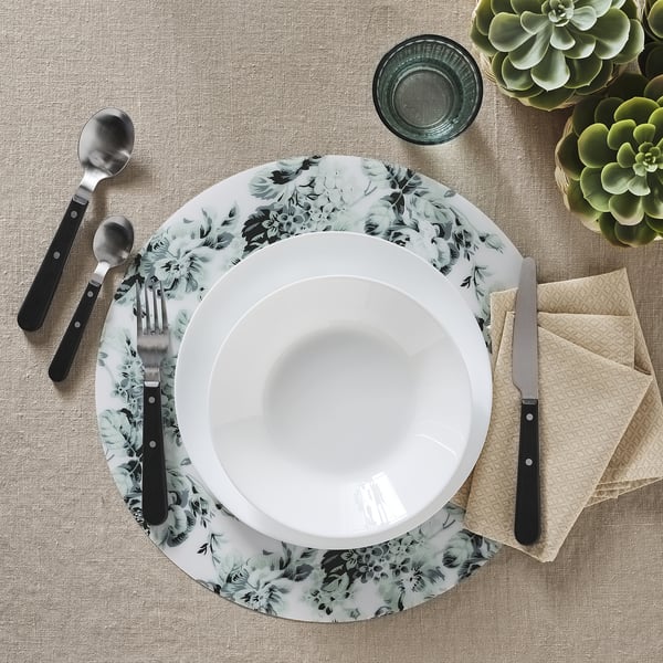 A table setting with white bowl, black-handled cutlery, floral plate, beige napkin, and glass on a woven placemat. Dishwasher and microwave-safe.