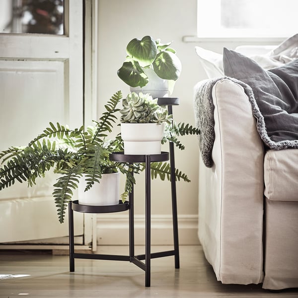 Modern black metal plant stand with three white pots holds various leafy plants, placed beside a cream sofa with blankets. Indoor home decor.