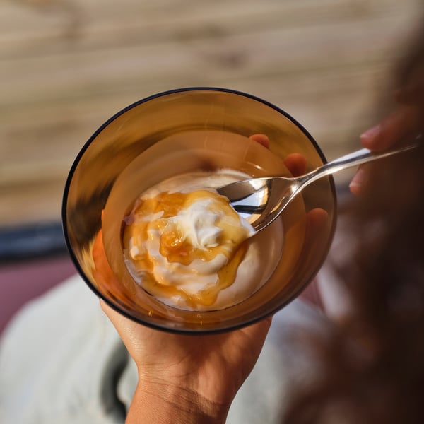 A hand holds a glass bowl filled with yoghurt and mango sauce. A spoon mixes the contents. The bowls subtle pattern highlights its handcrafted design.