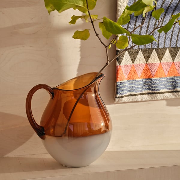 Ommjange amber glass jug with handle, round base, sits on wooden floor, beside plant.