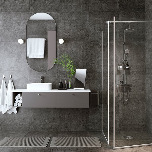 Modern bathroom with grey floating vanity, white sink, round mirror, glass shower door, and towels.