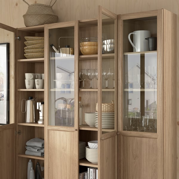 Wooden cabinet with glass doors showcasing dishes, glasses, and mugs. Shelves organised with baskets and stacks of plates and bowls.