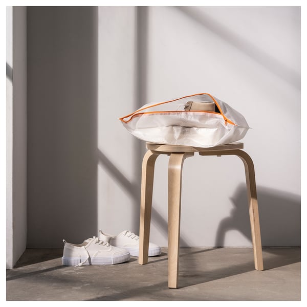 A light wood stool with a white mesh bag featuring orange trim sits against a white wall. White trainers are on the floor beside it.