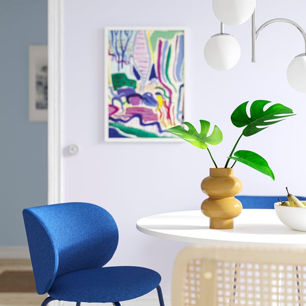 Modern dining area with yellow-beige vase, green plant, blue chair, and colourful painting.