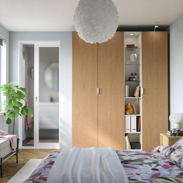 Modern bedroom with tall brown PAX wardrobe, sliding glass doors, patterned bedspread, plant, and pendant light.
