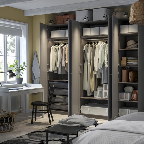Modern bedroom with an open wardrobe displaying clothes, folded shirts, and storage bins. A desk with a chair and a plant is nearby.