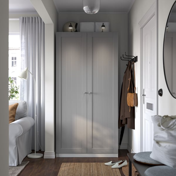 Modern grey PAX wardrobe with sleek handles stands by a white door with hooks, coat, and bag. Next to a sofa, lamp, and window with sheer curtains.