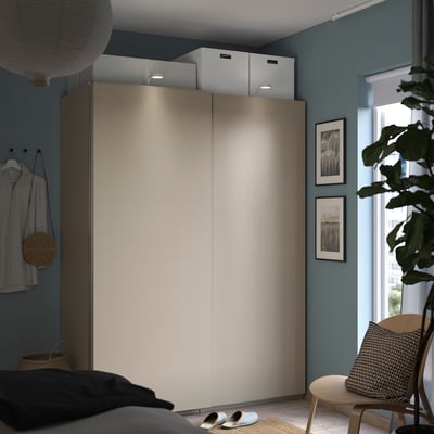 Modern bedroom with PAX beige wardrobe, blue walls, hanging shirt, window with plants, and bed with dark blanket.