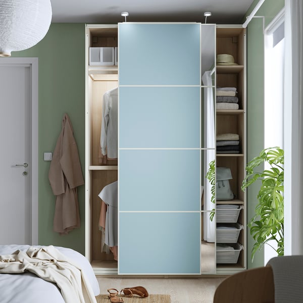 A bedroom showcasing a PAX wardrobe with sliding blue doors, revealing clothes hangers, shelves with folded towels, and storage bins.