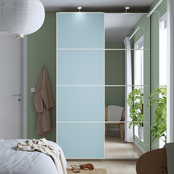 Modern bedroom with large light blue PAX wardrobe, sliding mirrors, & muted green walls. Includes white bed, potted plant, & wooden chair.