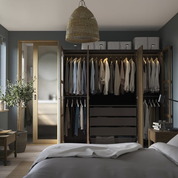 Bedroom with open wardrobe displaying clothes, drawers, and accessories on dark walls. Grey bed with white sheets in foreground.
