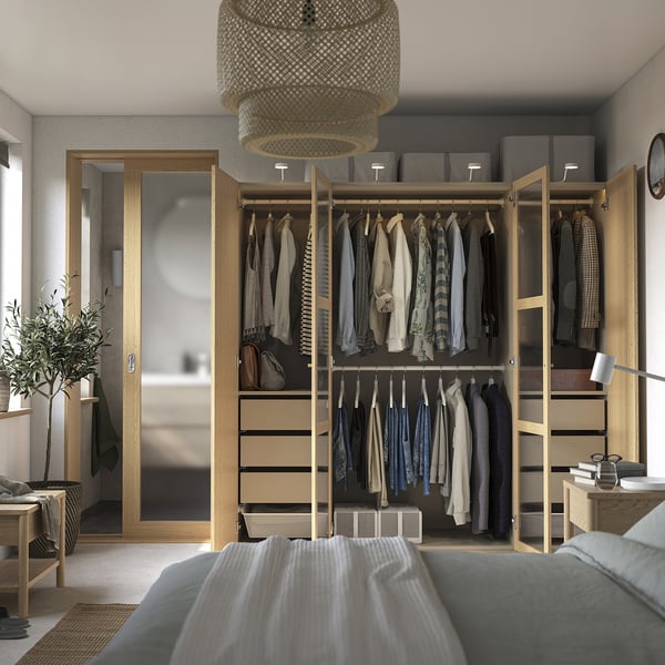 Bedroom with open wooden closet showing organised clothes, various drawers, and simple decor. Ceiling light fixture, mirror, and table lamp visible.