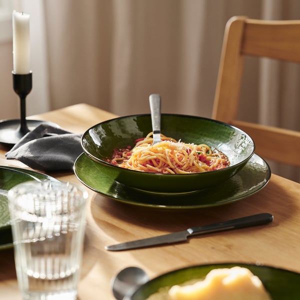 Green ceramic bowl holds pasta with fork; placed on wooden table with glass, knife, and chair.